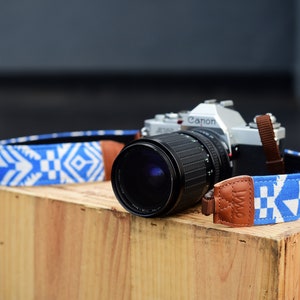May include: A silver Canon camera with a black lens and a blue and white patterned camera strap. The strap has a brown leather accent.