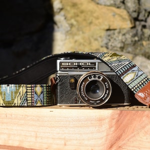 May include: A vintage black camera with a patterned strap rests on a wooden surface. The strap features a colorful geometric design with brown leather accents. The camera has the word "SOKOL" on it.