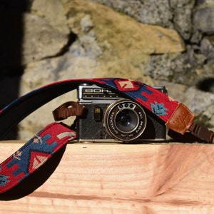 May include: A vintage black camera with a patterned red, blue, and tan camera strap. The camera strap is made of woven fabric and has leather ends. The camera is resting on a wooden surface.