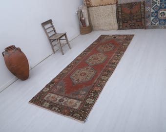Könnte beinhalten: Ein langer, rechteckiger Vintage-Teppich mit rotem Feld und komplizierten geometrischen Mustern in Beige, Braun und Grün. Der Teppich liegt auf einem weißen Boden, mit einem Holzstuhl und einer großen Terrakotta-Vase im Hintergrund.