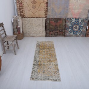 May include: A rectangular, distressed, vintage rug with a yellow and beige floral pattern. The rug is placed on a white floor, with a wooden chair and a large terracotta pot in the background. The rug measures approximately 60 x 180 cm.