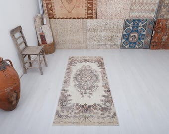 May include: A rectangular, cream-colored rug with a central medallion design and floral border. The rug features a faded, vintage aesthetic with brown and beige tones. The rug is placed on a white wooden floor.