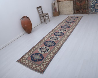 Puede incluir: Una alfombra larga y rectangular con un patrón repetitivo de formas geométricas azules y crema sobre un fondo rosa. La alfombra tiene un borde crema con un diseño rojo y marrón. Un jarrón de terracota y una silla de madera están en el fondo.