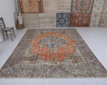 Puede incluir: Gran alfombra rectangular con un patrón intrincado. La alfombra presenta un medallón central en tonos naranja y azul, rodeado por un borde de tonos más oscuros. La alfombra está colocada sobre un suelo blanco, con una silla de madera y otras alfombras en el fondo.
