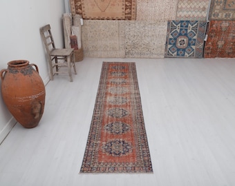 May include: An antique runner rug with a distressed, vintage look. The rug features a central design in shades of orange, blue, and cream, with a dark border. The rug is displayed on a white floor.
