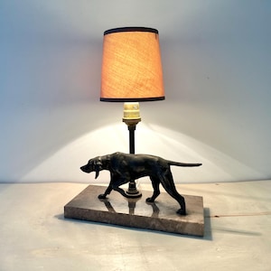Op de afbeelding: Een decoratieve tafellamp met een zwart hondbeeldje op een rechthoekige basis. De lamp heeft een bruine lampenkap met een zwarte rand en een messingkleurige lamphouder. De lamp is ontworpen om op een tafel of ander plat oppervlak te staan.