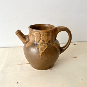 Puede incluir: Una jarra de cerámica marrón con un esmalte marrón claro y un patrón moteado. La jarra tiene un asa y un pico.