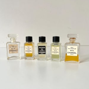 Pode incluir: Cinco frascos de perfume Chanel vintage. Os frascos são de vidro transparente com rótulos retangulares e tampas pretas ou brancas. Os frascos contêm vários perfumes coloridos, incluindo amarelo e verde. Os rótulos exibem a marca Chanel e os nomes dos perfumes.