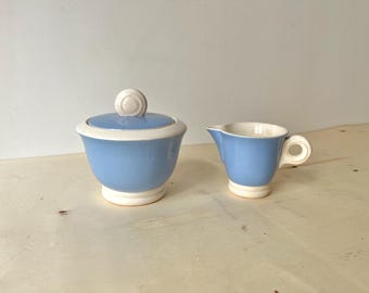 Może przedstawiać: Jasnoniebieska ceramiczna cukiernica z pokrywką i pasującą śmietanką. Cukiernica ma białą obwódkę i podstawę oraz białą gałkę na pokrywce. Śmietanka ma uchwyt i dzióbek. Zestaw jest prawdopodobnie w stylu vintage.