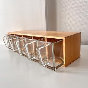 Puede incluir: Un especiero de madera con seis recipientes de vidrio transparente, cada uno con un asa. El estante es de madera clara y se encuentra sobre una superficie blanca. Los recipientes de vidrio son rectangulares.