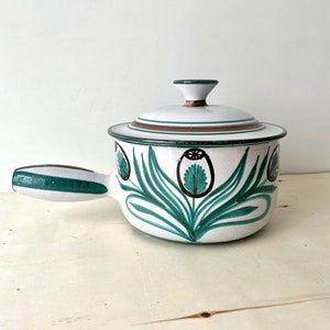 Op de afbeelding: Witte keramische pot met een groen en bruin bloemenmotief. De pot heeft een deksel en een handvat.