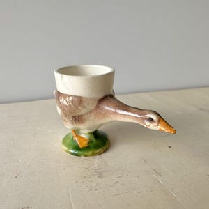 以下が含まれることがあります： クリーム色の体、茶色の翼のディテール、オレンジ色のくちばしを持つ、ガチョウの形をした陶器製の卵立て。ガチョウは小さな緑色の台座の上に立っており、長い首が外側に伸びて容器の側面を形成している機能的な器である。