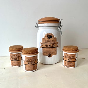 Puede incluir: Cuatro tarros de cerámica blanca con tapas de corcho y etiquetas marrones. Las etiquetas dicen "Terracotta from Tuscany" y tienen diferentes nombres para cada tarro: "Pickles", "Pepper", "Spices" y "Salt".