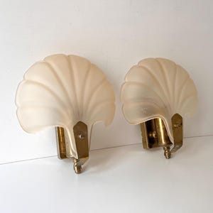 Könnte beinhalten: Zwei Vintage-Wandleuchten mit Muscheldesign. Die Leuchten haben cremefarbene, satinierte Glasschirme und Messingfarbene Metallbeschläge. Die fächerförmigen Schirme sind an einer weißen Wand montiert.