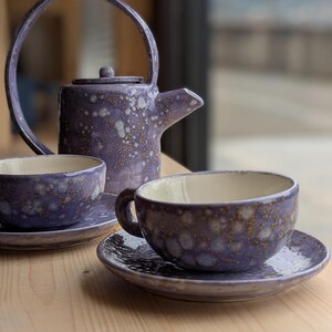 Può includere: Servizio da tè in ceramica viola con smalto maculato. Il set comprende una teiera con manico curvo, due tazze e due piattini. L'interno delle tazze è color crema. Il set è esposto su una superficie di legno.