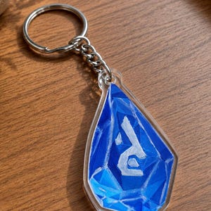 Peut inclure: Un porte-clés bleu et blanc avec un motif géométrique. Le porte-clés a une chaîne et un anneau argentés. La partie bleue du porte-clés comporte la lettre blanche "R" au centre.
