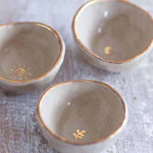 Może przedstawiać: Trzy małe, ręcznie robione ceramiczne miseczki w neutralnym beżowym kolorze ze złotymi obwódkami. Każda miseczka ma złoty wzór: słońce, półksiężyc i liść. Miseczki ułożone są na jasnej, teksturowanej powierzchni.