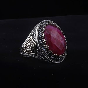 Peut inclure: Une bague en argent avec un motif orné et une grande pierre précieuse de rubis de forme ovale entourée de petites pierres précieuses noires.