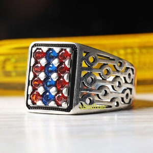 Könnte beinhalten: Ein silberner Ring mit einem quadratischen Design, das mit roten und blauen Edelsteinen besetzt ist. Der Ring hat ein filigranes durchbrochenes Muster auf der Band. Der Text "sbisilver" ist auf dem Ring sichtbar.