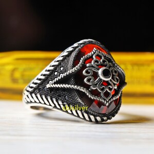 May include: A silver ring featuring a large red gemstone set within an intricate filigree design. The ring is adorned with smaller black gemstones.