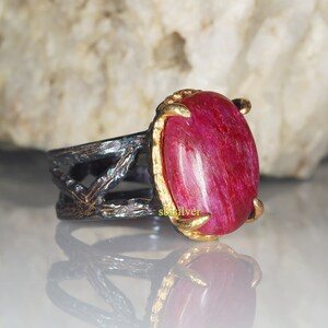 May include: A silver ring with a red gemstone set in a gold claw setting. The band of the ring is black and has a textured design.