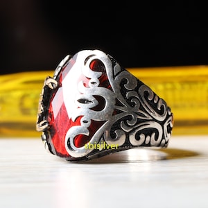 May include: A silver ring featuring a red gemstone set within a filigree design. The ring is engraved with the text "sbisilver".