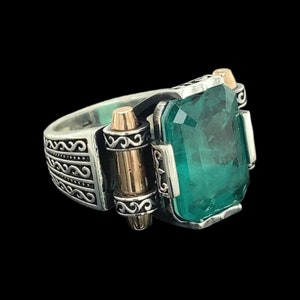 May include: A silver ring featuring a large green gemstone and gold accents. The ring has a detailed design with intricate patterns.
