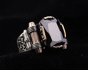 May include: A silver ring with a black gemstone and intricate details. The ring has a decorative band with a scroll design and a raised section with a black stone.