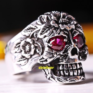 May include: A silver skull ring with intricate floral details and two large ruby red stones for eyes. The ring is a statement piece with a gothic and edgy style.
