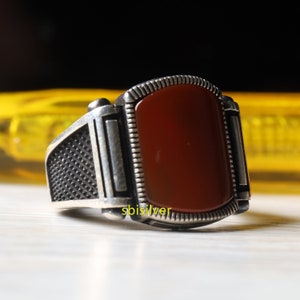 May include: A silver ring with a dark red stone set in a rectangular shape. The ring has a textured band and a decorative border around the stone.