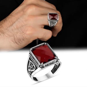 Puede incluir: Un anillo de plata con una gran gema rectangular facetada de color rojo intenso. El anillo tiene adornos de volutas en los laterales y un acabado pulido. El anillo se muestra en una mano y también aislado.