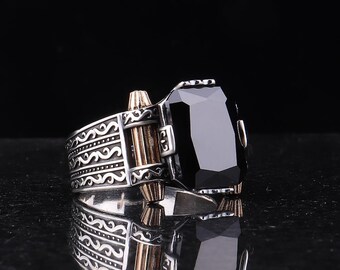 May include: A silver ring with a large, rectangular black gemstone. The ring features intricate scrollwork and gold-colored accents. The ring is displayed on a reflective surface against a black background.