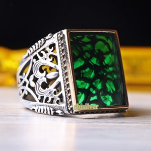 Könnte beinhalten: Ein silberner Ring mit einem verzierten Design und einem großen, rechteckigen grünen Edelstein. Der Ring ist mit einem goldenen Band besetzt.