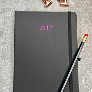 Könnte beinhalten: Ein schwarzes Notizbuch mit einem schwarzen Gummibandverschluss und dem Wort "WTF" in Pink auf dem Einband. Ein Bleistift mit rotem Radiergummi liegt auf dem Notizbuch.