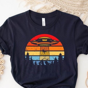 May include: A navy blue t-shirt with a retro-style graphic of a UFO flying over a sunset. The graphic features a colorful sunset with a red, orange, yellow, and blue color scheme. The UFO is white and has a red light on the bottom.
