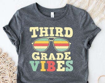 May include: A dark grey t-shirt with the text "THIRD GRADE VIBES" in a retro font. The text is in colourful letters with a pair of sunglasses with colourful stripes above the text.