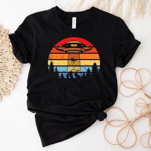 May include: Black t-shirt with a retro sunset graphic featuring a flying saucer and trees.