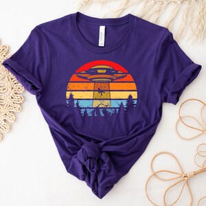 May include: Purple t-shirt with a retro-style graphic of a UFO flying over a sunset with trees in the foreground.