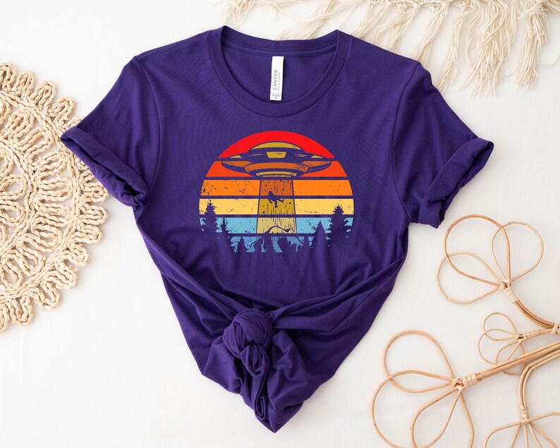 May include: Purple t-shirt with a retro-style graphic of a UFO flying over a sunset with trees in the foreground.
