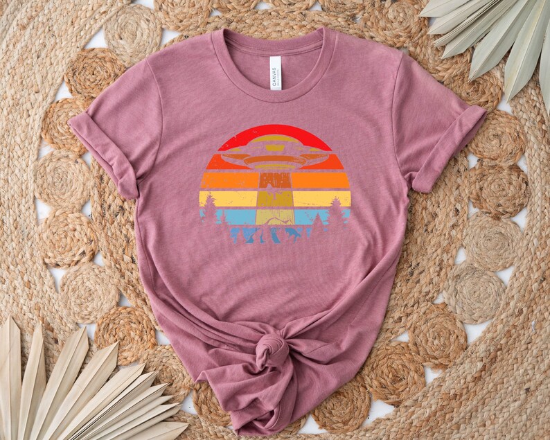 May include: A mauve t-shirt with a vintage-style graphic of a UFO flying over a sunset. The graphic is in shades of orange, yellow, blue, and green.