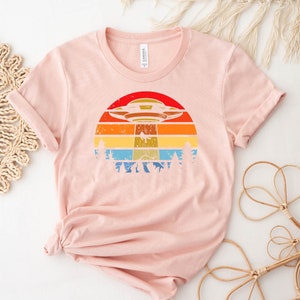 May include: A pink t-shirt with a retro-style graphic of a UFO flying over a sunset with trees in the foreground.