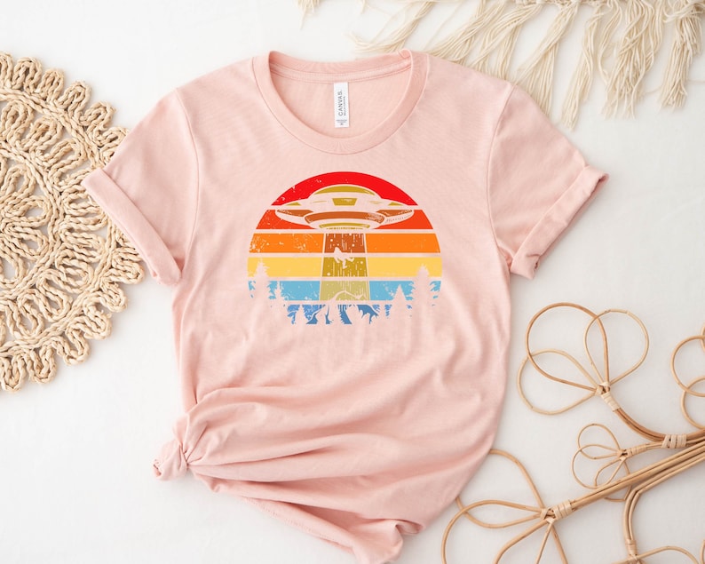May include: A pink t-shirt with a retro-style graphic of a UFO flying over a sunset with trees in the foreground.