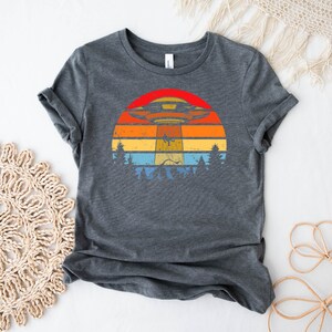 May include: A dark gray t-shirt with a retro-style graphic of a UFO flying over a sunset with trees in the foreground.