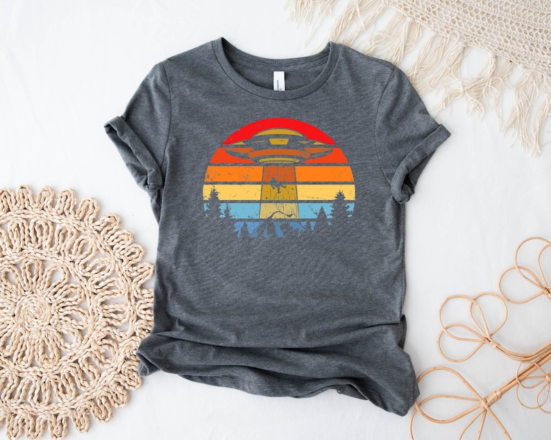 May include: A dark gray t-shirt with a retro-style graphic of a UFO flying over a sunset with trees in the foreground.