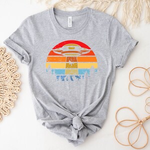 May include: A gray t-shirt with a retro-style graphic of a UFO flying over a sunset. The graphic features a colorful striped sunset with a dark blue sky and a white UFO.