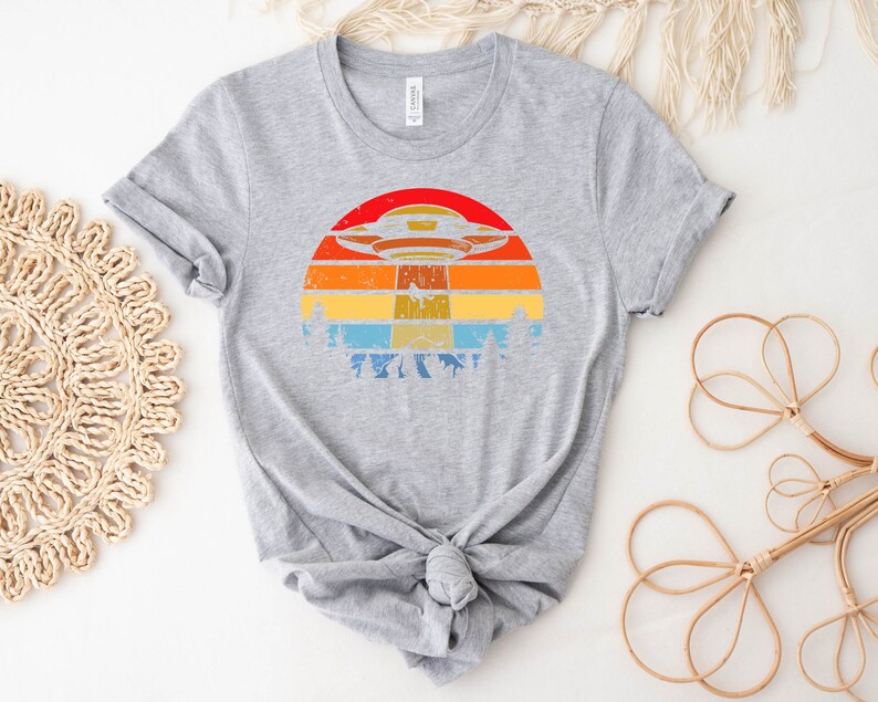 May include: A gray t-shirt with a retro-style graphic of a UFO flying over a sunset. The graphic features a colorful striped sunset with a dark blue sky and a white UFO.