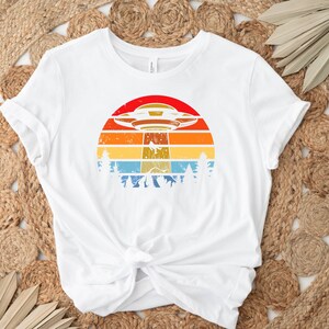 May include: White t-shirt with a retro-style graphic of a UFO flying over a sunset with trees in the foreground.