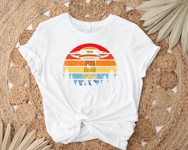 May include: White t-shirt with a retro-style graphic of a UFO flying over a sunset with trees in the foreground.