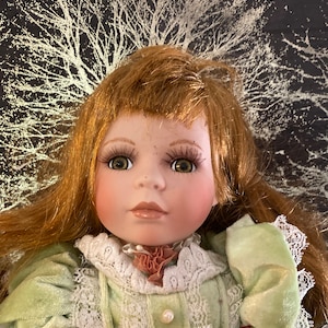 May include: A porcelain doll with long, auburn hair wearing a green velvet dress with white lace trim and a pink flower accent. The doll has green eyes and a delicate facial expression. The background features a gold and white tree design.