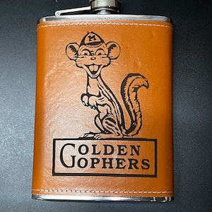 May include: A brown leather-covered flask with a silver top and a black and white image of a gopher wearing a hat. The text "GOLDEN GOPHERS" is engraved below the image.